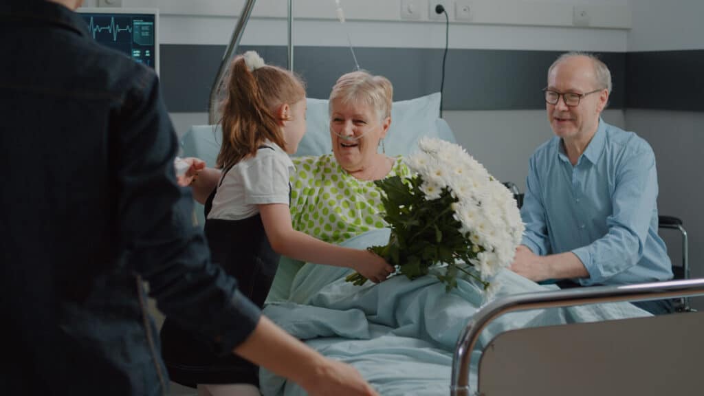 Old patient receiving visit from child and mother in hospital ward. Kid with flowers running to hug sick woman in bed, visiting grandma to give comfort and help with recovery. Family at clinic
