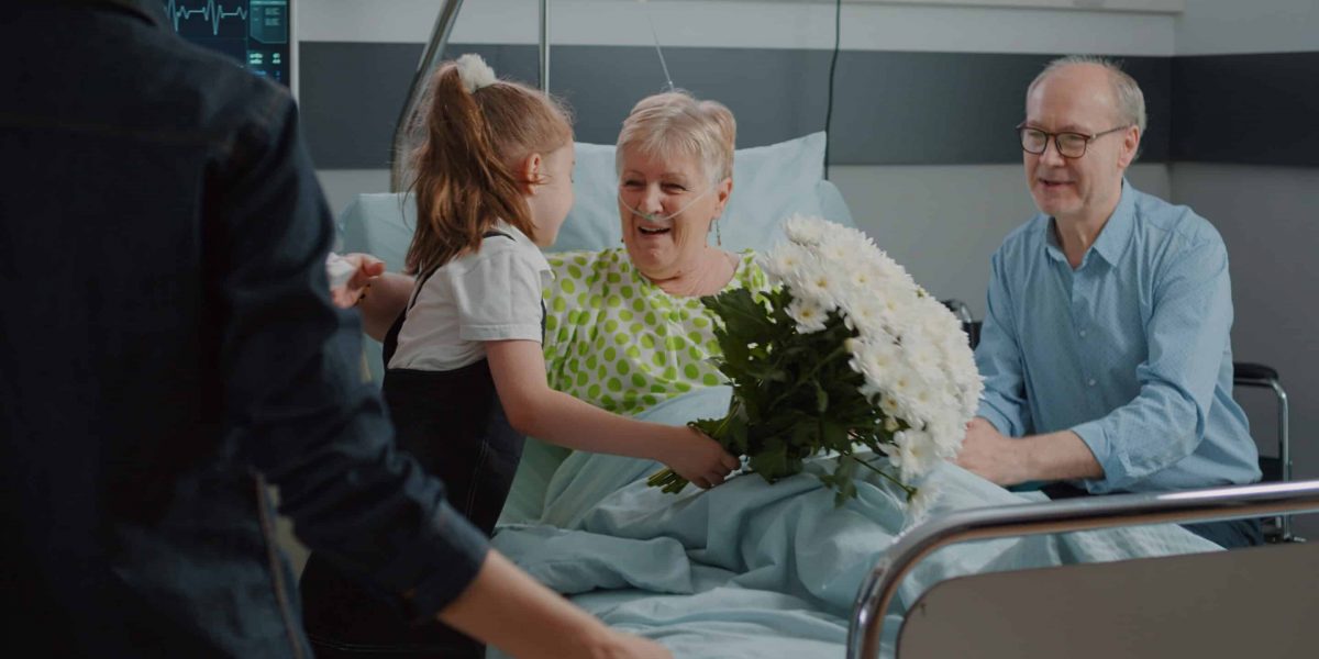 Old patient receiving visit from child and mother in hospital ward. Kid with flowers running to hug sick woman in bed, visiting grandma to give comfort and help with recovery. Family at clinic